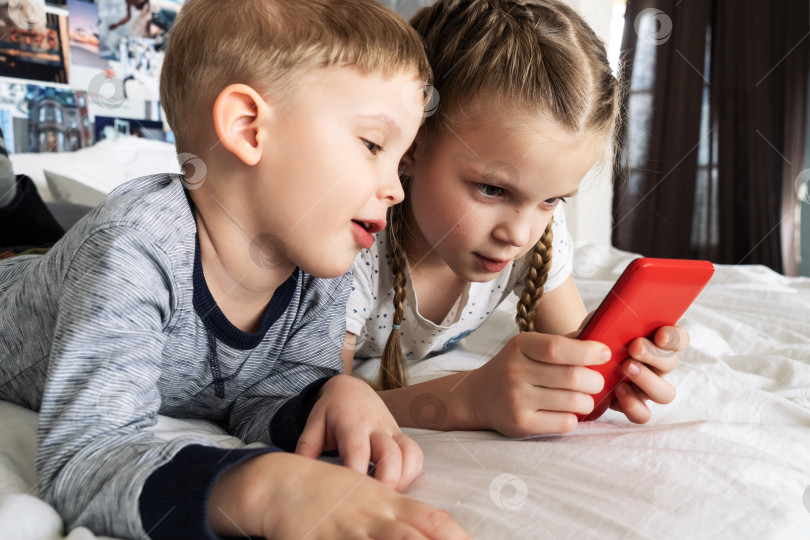 Скачать Детские гаджеты: мальчик скептически заглядывает в смартфон к девочке. Оба лежат на кровати. фотосток Ozero
