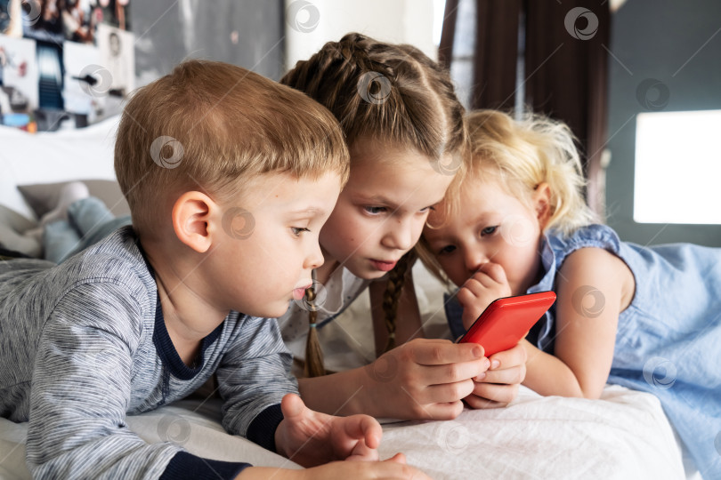 Скачать Поколение ZD: Дети лежат на кровати и смотрят в красный смартфон старшей девочки. фотосток Ozero