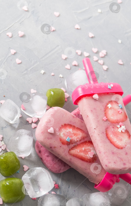 Скачать Летняя жара: освежающее розовое фруктовое мороженое с кусочками клубники в окружении прозрачных и зеленых кубиков льда на сером столе. фотосток Ozero