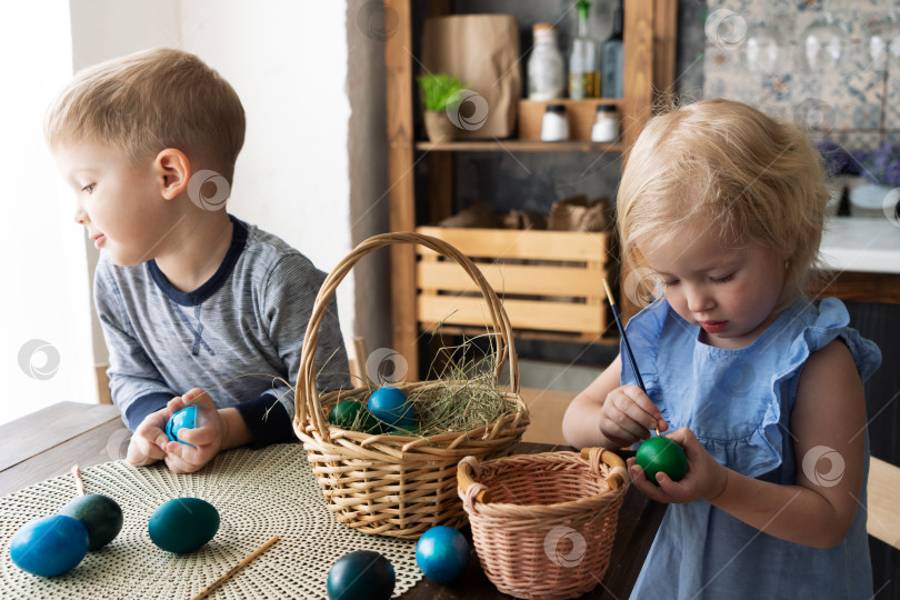 Скачать Праздник Пасхи: Девочка и мальчик рисуют кисточкой яйца на кухне. фотосток Ozero