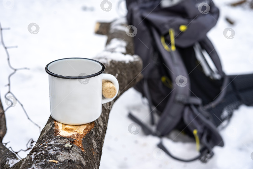 Скачать Горячие напитки в зимнем походе: Белая железная эмалированная кружка и рюкзак на заднем плане. фотосток Ozero