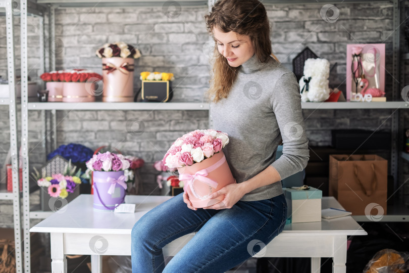 Скачать Девушка-флорист сидит на столе в мастерской и держит в руках круглую розовую коробку с букетом роз. фотосток Ozero