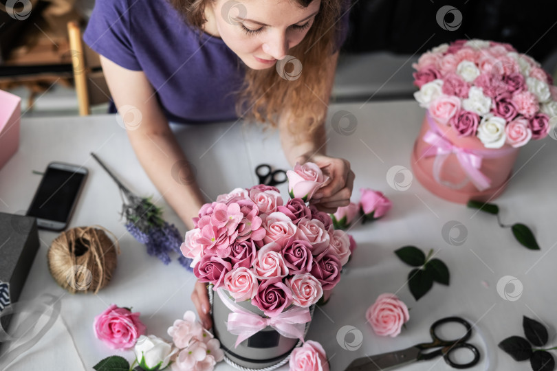 Скачать Цветочный магазин: девушка-флорист собирает букет розовых роз в круглую коробку. Светлые вьющиеся волосы на сером фоне. фотосток Ozero