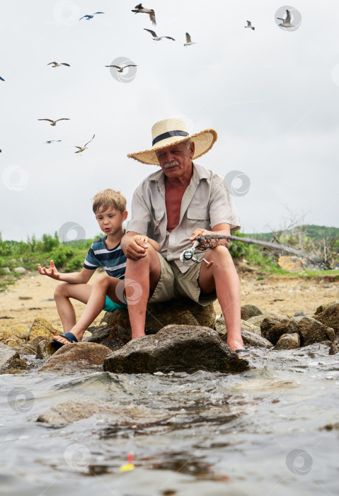 Скачать Дедушка-рыбак в соломенной шляпе и маленький внук мужского пола, наслаждающиеся досугом, пользуются удочкой фотосток Ozero