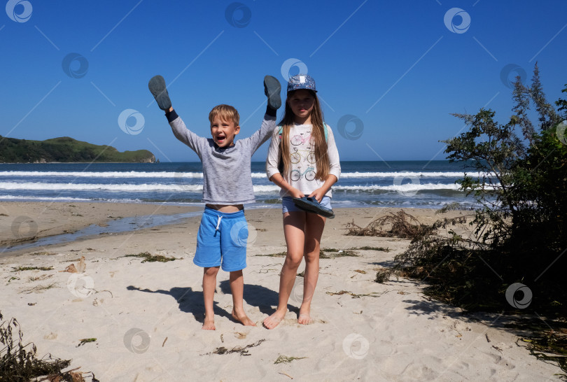Скачать Счастливые маленькие дети играют в туфлях на руках, позируя на прекрасном песчаном пляже с морским пейзажем, наслаждаясь отдыхом фотосток Ozero