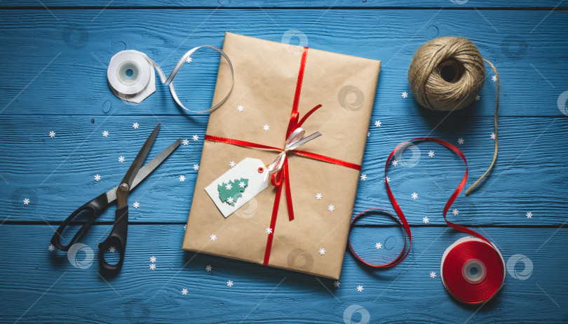 Скачать Подарочная упаковка из бумаги ручной работы и красной ленты на синих деревянных досках с белыми снежинками фотосток Ozero