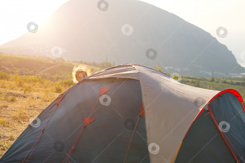 Скачать Веселый ребенок выглядывает из-за туристической палатки в горах с панорамным видом на рассвете. Внутренний туризм, активная летняя поездка, семейные приключения. Экотуризм, кемпинг, спортивные походы в горы. фотосток Ozero
