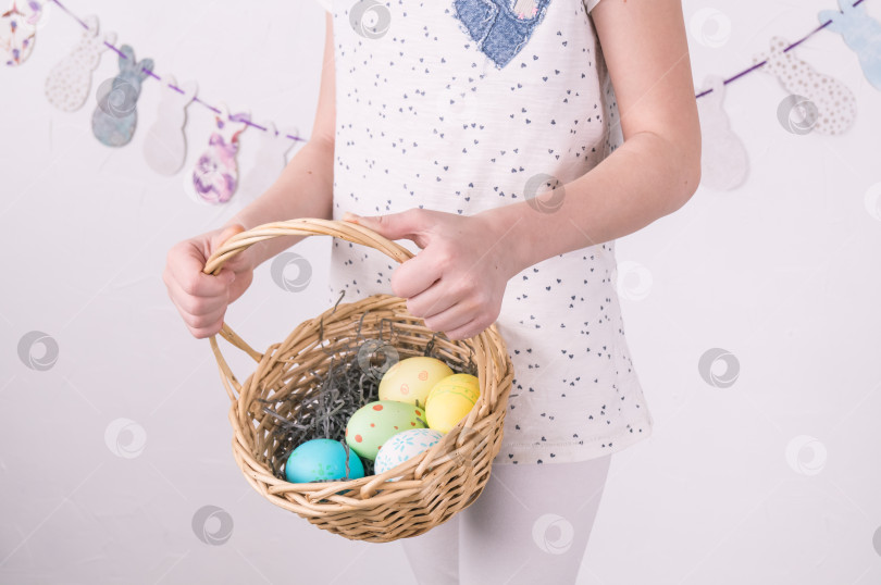 Скачать Поздравления со счастливой Пасхой: девушка держит в руках корзину с крашеными яйцами у белой стены. фотосток Ozero