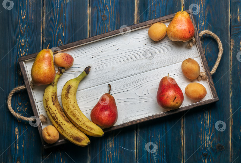 Скачать Свежие фрукты на белом деревянном подносе: бананы, груши, абрикосы. Сельская местность. Вид сверху. фотосток Ozero