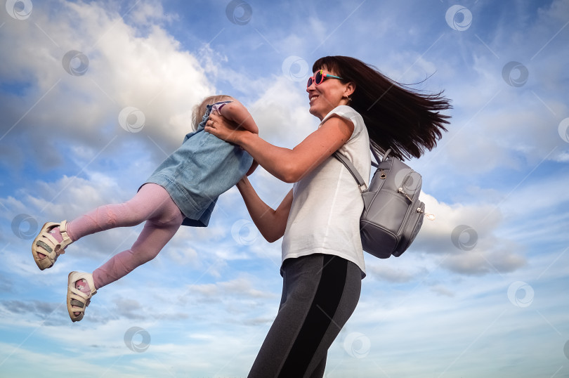 Скачать Семейные ценности: Мама в солнцезащитных очках кружится с маленькой дочкой в голубом платье на фоне неба и облаков. Настоящая семья. фотосток Ozero