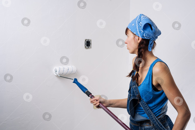 Скачать Девушка в синей одежде обливает валиком белую стену в комнате. фотосток Ozero