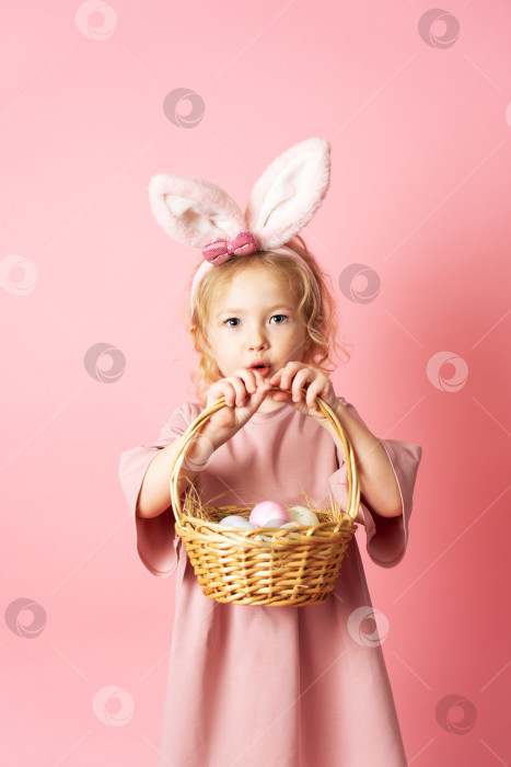 Скачать Счастливой Пасхи: маленькая девочка в розовом платье и с заячьими ушками держит в руках корзинку с разноцветными яйцами. фотосток Ozero