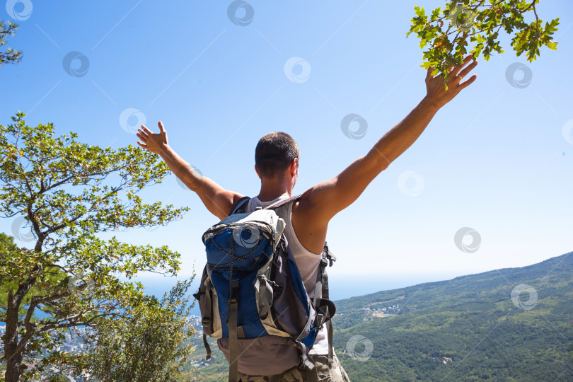 Скачать Мужчина-турист с рюкзаком на вершине горы с поднятыми руками смотрит на панораму побережья, города, радуется свободе. Путешествия, треккинг, пеший туризм, активный образ жизни, спортивный туризм, пешие прогулки. фотосток Ozero