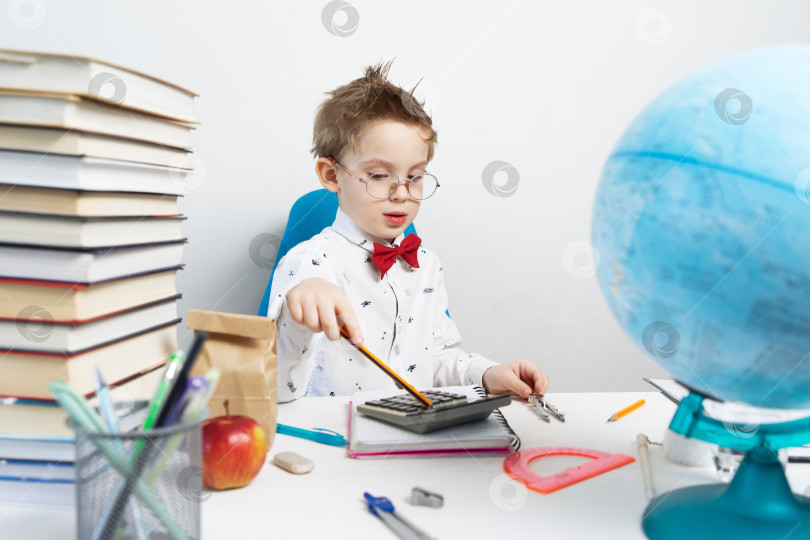 Скачать Возвращение в школу: маленький мальчик в рубашке и очках сидит за своим столом и делает вычисления на калькуляторе. фотосток Ozero