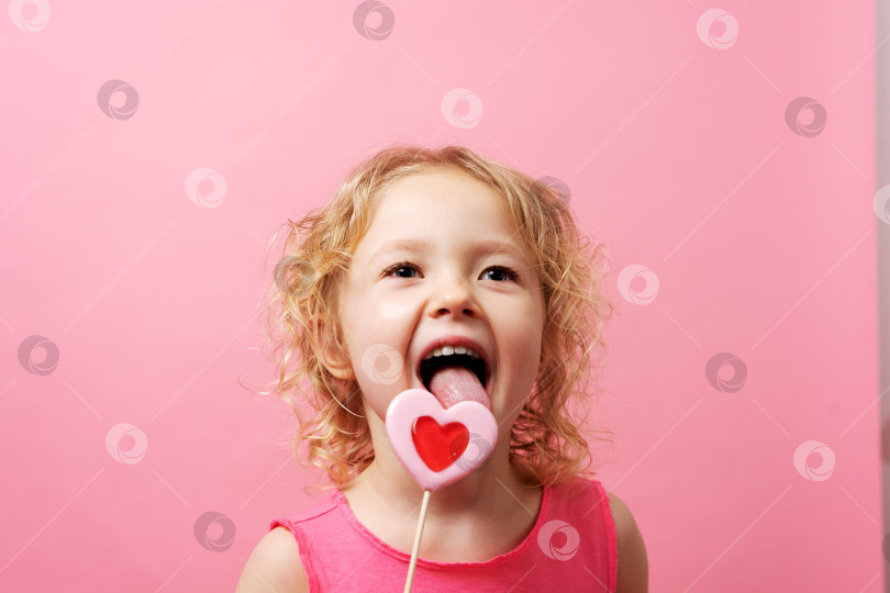 Скачать Маленькая девочка держит в руках большие леденцы в форме сердца на розовом фоне. фотосток Ozero