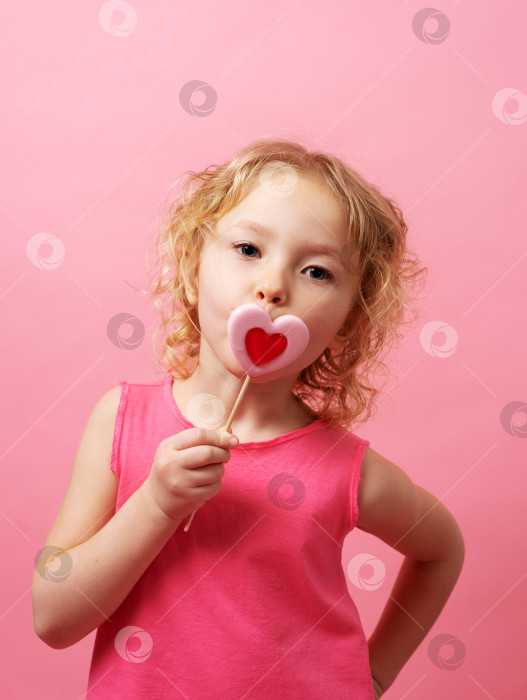 Скачать Маленькая девочка держит в руках большие леденцы в форме сердца на розовом фоне. фотосток Ozero