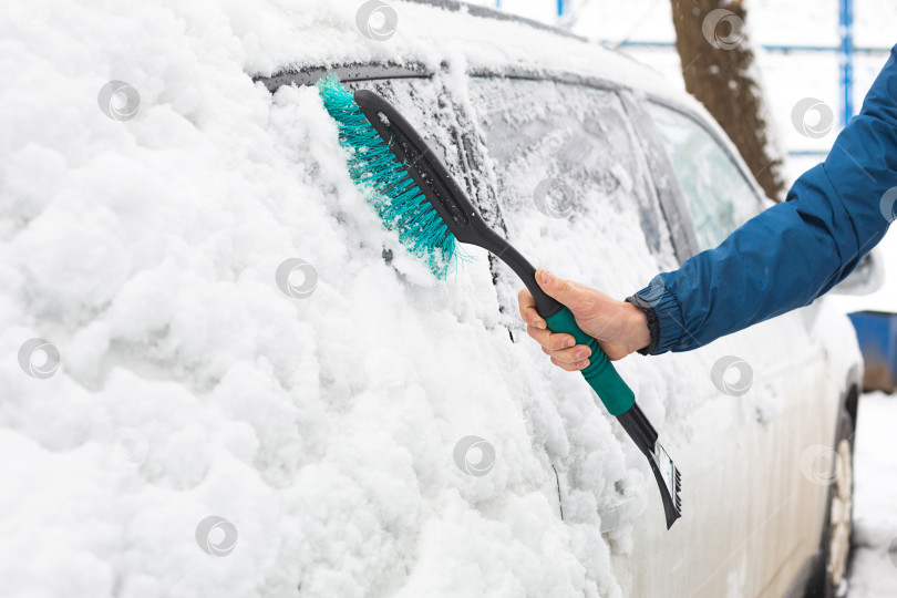 Скачать Мужчина счищает снег с машины после снегопада. Рука в синей куртке с автомобильной метлой на белом теле. Зимние погодные условия фотосток Ozero