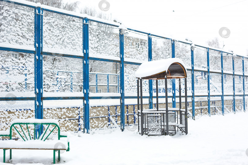 Скачать Заснеженный двор жилого квартала: высокий забор, спортивная баскетбольная площадка, скамейка, беседка. Зимняя снежная атмосфера фотосток Ozero