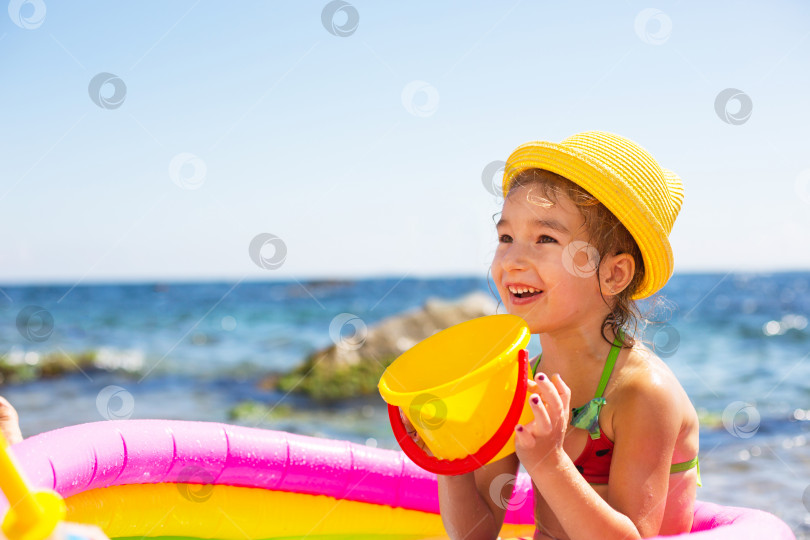 Скачать Девушка в желтой соломенной шляпе играет с ветром, водой и диспенсером для воды в надувном бассейне на пляже. Несмываемые средства для защиты детской кожи от солнца, солнечных ожогов. курорт на берегу моря. фотосток Ozero
