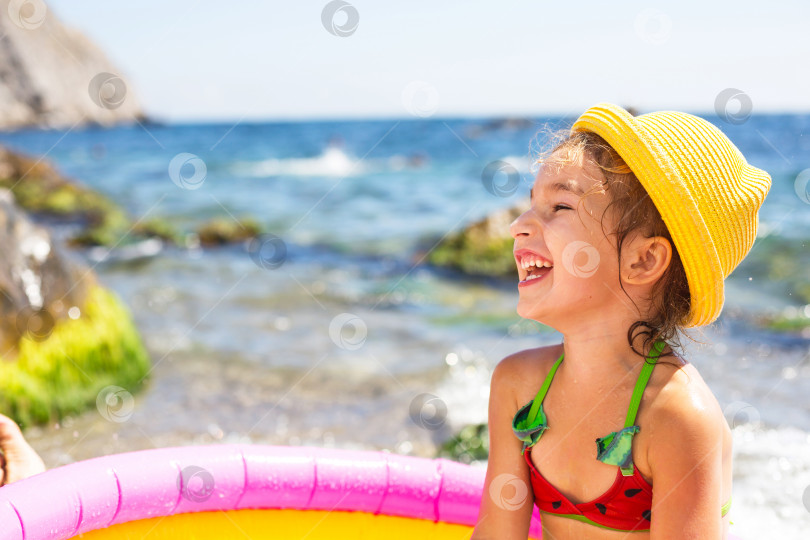 Скачать Девушка в желтой соломенной шляпе играет на открытом воздухе у моря, в воде с ведром в надувном бассейне на пляже. Несмываемые средства для защиты детской кожи от солнца, солнечных ожогов. курорт на берегу океана. фотосток Ozero