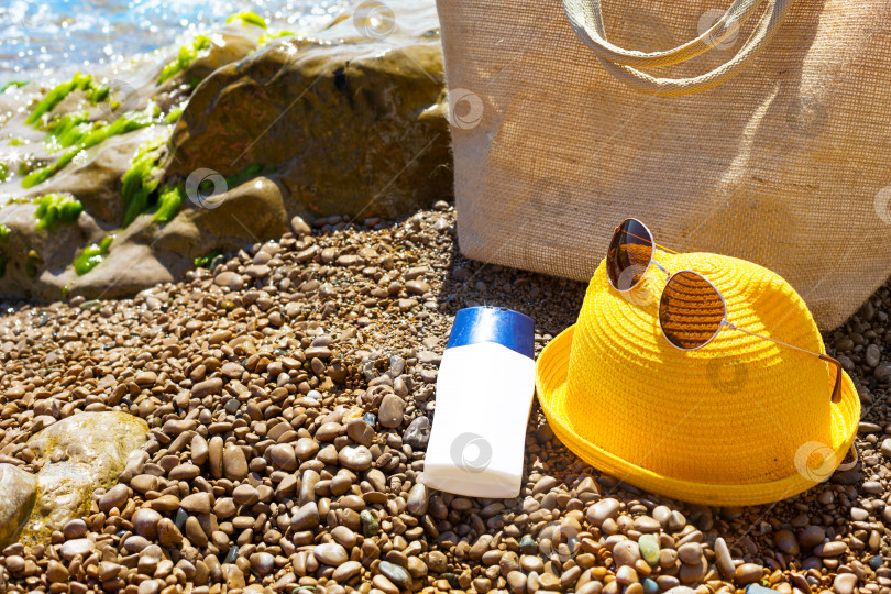 Скачать Пляжная сумка с белым тюбиком солнцезащитного крема на галечном пляже у моря. Путешествия, пляжный отдых на курорте, очки, желтая шляпа. Защита кожи от ультрафиолетовых лучей, масло для загара. Скопируйте пространство, смоделируйте. фотосток Ozero