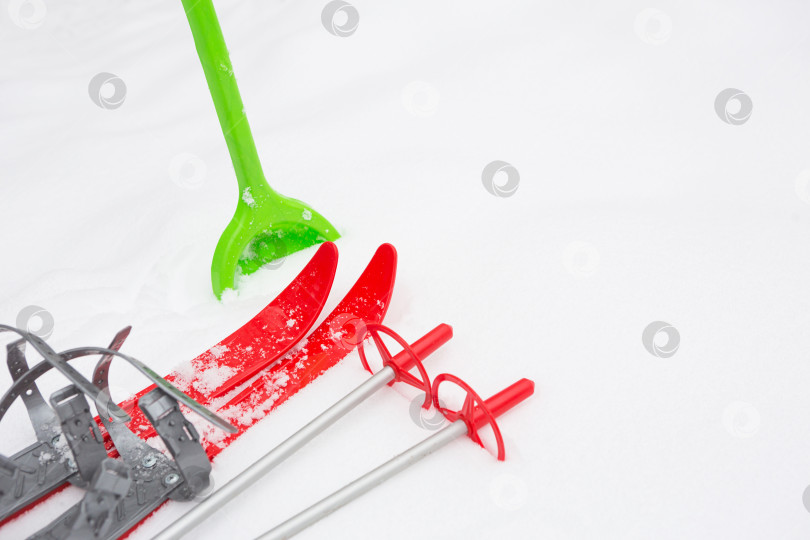 Скачать Детские красные лыжи с палками и зеленая лопата для уборки снега-макет на снегу. Зимние развлечения на свежем воздухе, семейные развлечения. Белый естественный морозный фон. Пространство для копирования фотосток Ozero