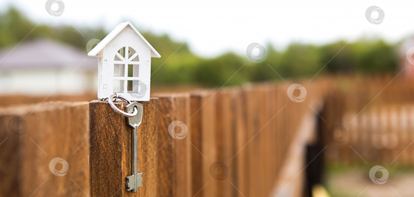 Скачать Маленькая фигурка белого деревянного дома на заборе с ключом от жилья на фоне коттеджей. Строительство, дизайн, проект, переезд в новый дом, ипотека, аренда и покупка недвижимости. Пространство для копирования фотосток Ozero