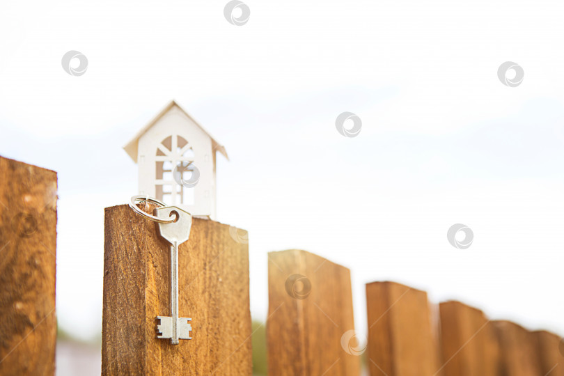 Скачать Маленькая фигурка белого деревянного дома на заборе с ключом от жилья на фоне коттеджей. Строительство, дизайн, проектирование, переезд в новый дом, ипотека, аренда и покупка недвижимости. Пространство для копирования фотосток Ozero