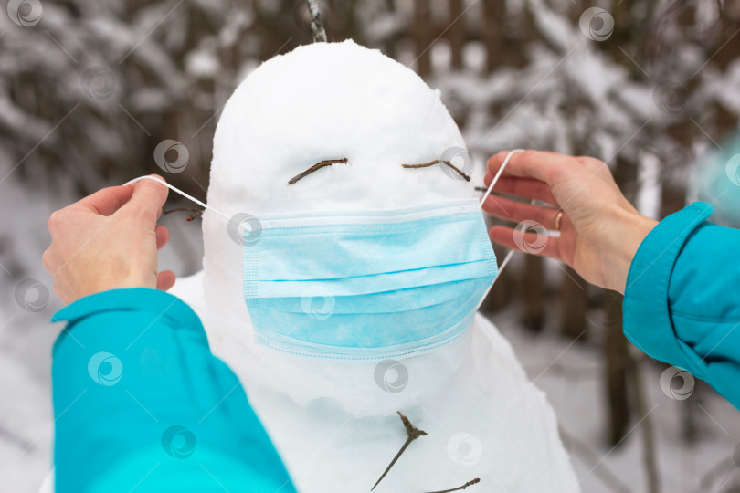 Скачать Снеговик в медицинской маске - новая реальность, защита от болезней, инфекций, жизнь в условиях эпидемии covid. Женские руки надевают маску снеговика. Зимние семейные мероприятия на свежем воздухе фотосток Ozero