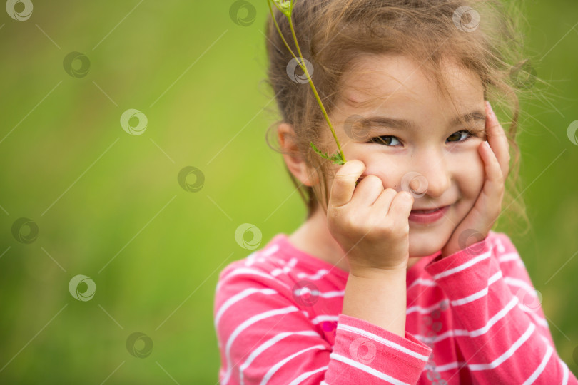 Скачать Маленькая девочка в футболке в коралловую полоску на зеленом фоне в поле закрывает лицо руками и лукаво улыбается. День защиты детей, счастливый ребенок, охрана окружающей среды и природопользования, средство от насекомых фотосток Ozero