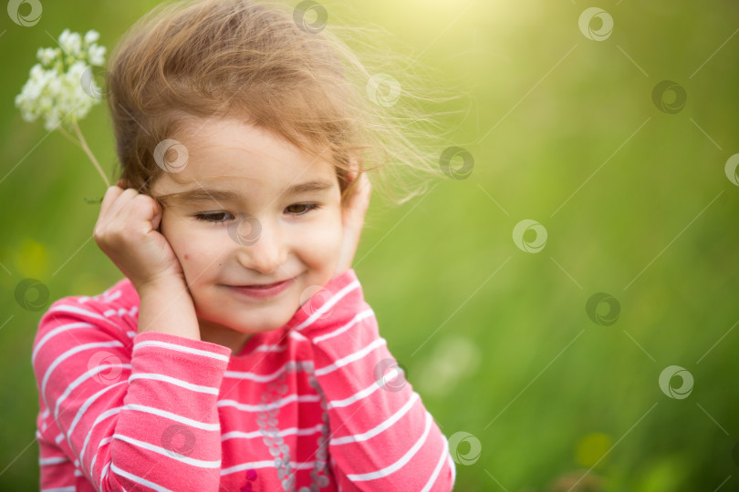 Скачать Маленькая девочка в футболке в коралловую полоску на зеленом фоне в поле держит лицо в ладонях и лукаво улыбается. День защиты детей, счастливый ребенок, охрана окружающей среды и природопользования, средство от насекомых фотосток Ozero