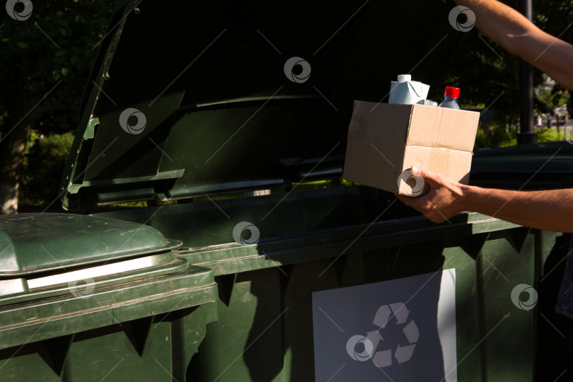 Скачать Картонная коробка с мусором в руках мужчины возле контейнеров для раздельного сбора отходов. Экологическая осведомленность населения, забота об окружающей среде, разделение: пластик, картон, упаковка. фотосток Ozero