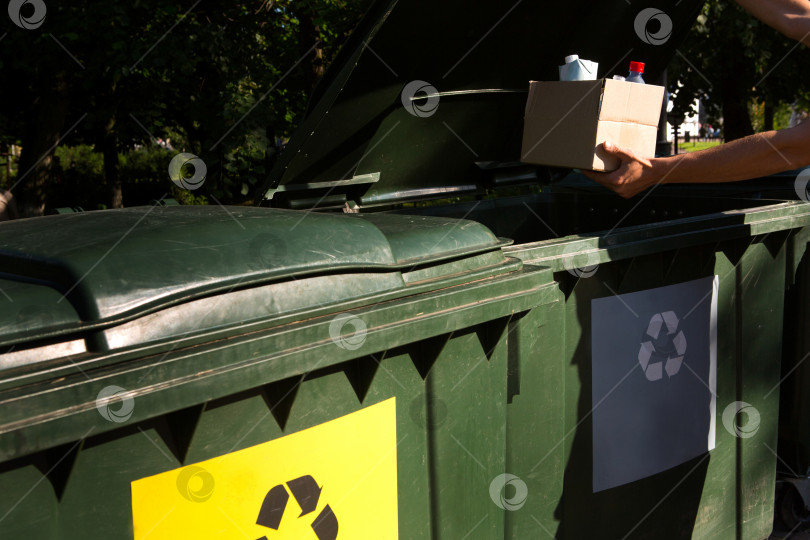 Скачать Картонная коробка с мусором в руках мужчины возле контейнеров для раздельного сбора отходов. Экологическое просвещение населения, забота об окружающей среде, разделение: пластика, картона, упаковки. фотосток Ozero