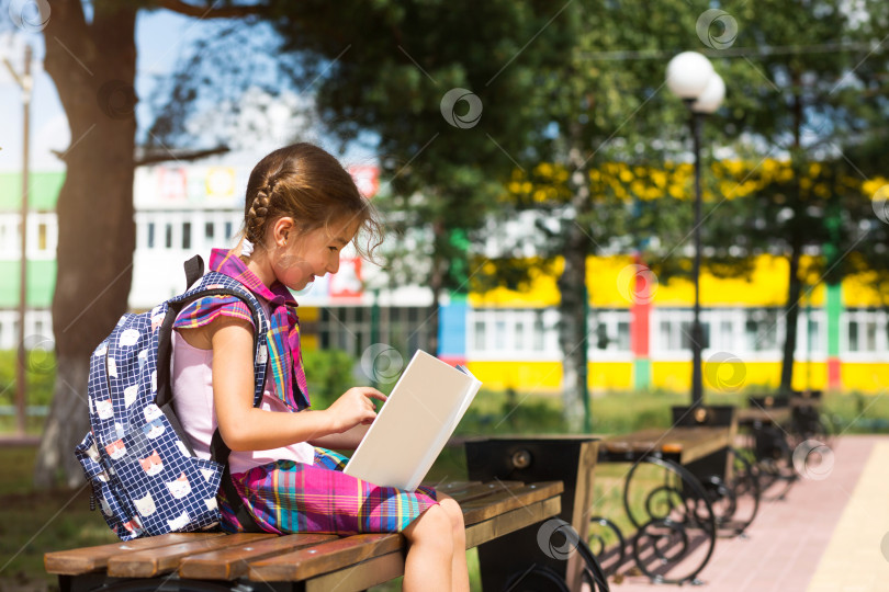 Скачать Девочка с рюкзаком сидит на скамейке и читает книгу возле школы. Возвращение в школу, расписание уроков, дневник с оценками. Образование, классы начальной школы, 1 сентября фотосток Ozero