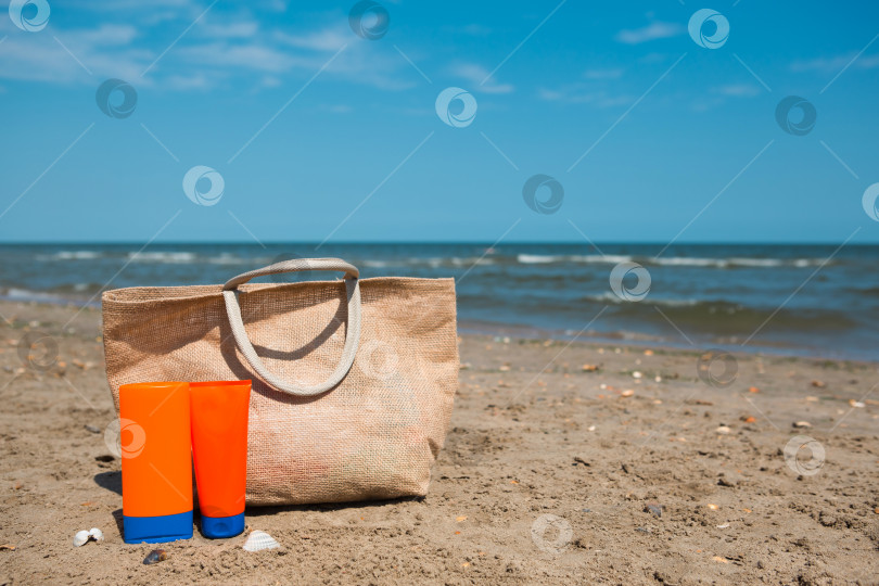 Скачать Оранжевый тюбик с макетом солнцезащитного крема, солнцезащитные очки и пляжная сумка стоят на песке у моря и покачиваются на ветру. Защита от ультрафиолета, пляжный отдых, путешествия фотосток Ozero