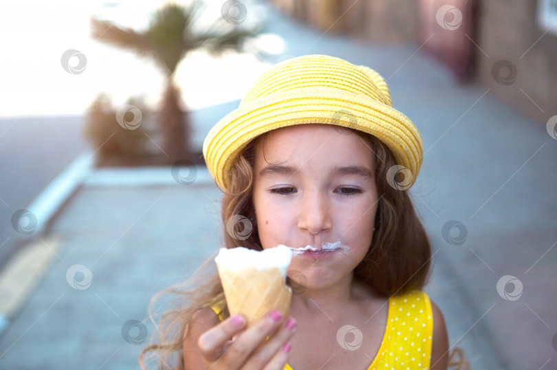 Скачать Летом ребенок с удовольствием ест вкусное мороженое на свежем воздухе, испачкав рот. Девушка в желтой шляпке и сарафане в жару на портрете крупным планом фотосток Ozero