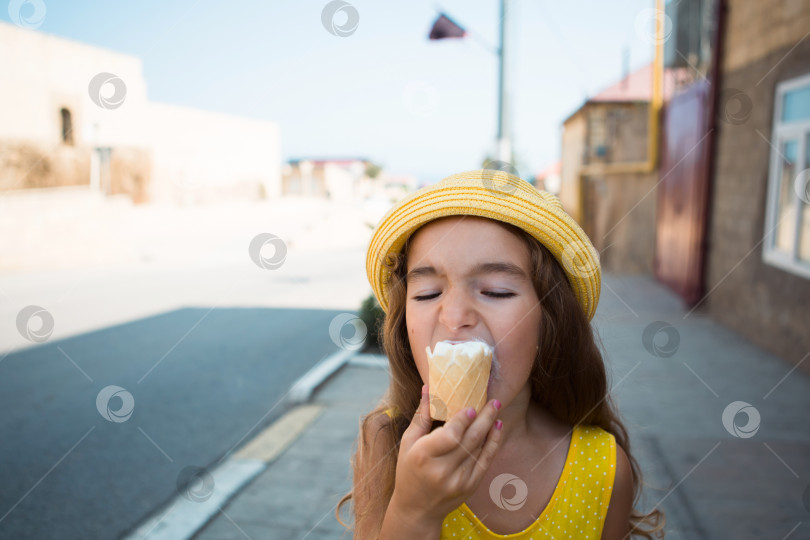 Скачать Летом ребенок с удовольствием ест вкусное мороженое на свежем воздухе, испачкав рот. Девушка в желтой шляпке и сарафане в жару на портрете крупным планом фотосток Ozero