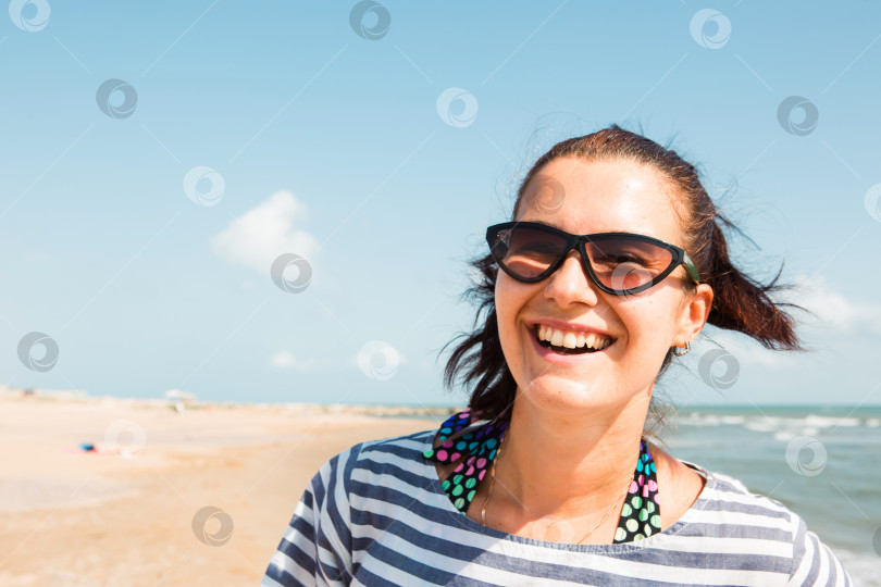 Скачать Портрет крупным планом счастливой брюнетки в полосатом платье и солнцезащитных очках стоит на берегу моря, смотрит на волны, смеется и улыбается. Туризм, пляжный отдых, бриз, путешествия, travel фотосток Ozero