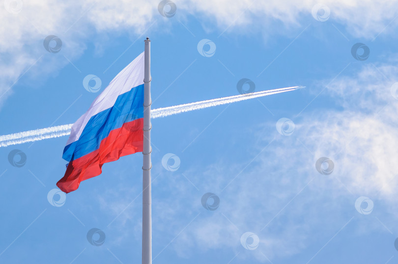 Скачать Флаг России на фоне голубого облачного неба с белым следом от самолета. фотосток Ozero