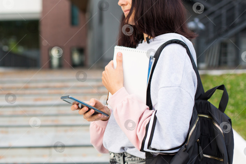 Скачать Студентка с черным рюкзаком держит смартфон и блокнот на пороге университета. Высшее образование, начало учебного года, возвращение в школу, гаджеты. Копировальное пространство фотосток Ozero