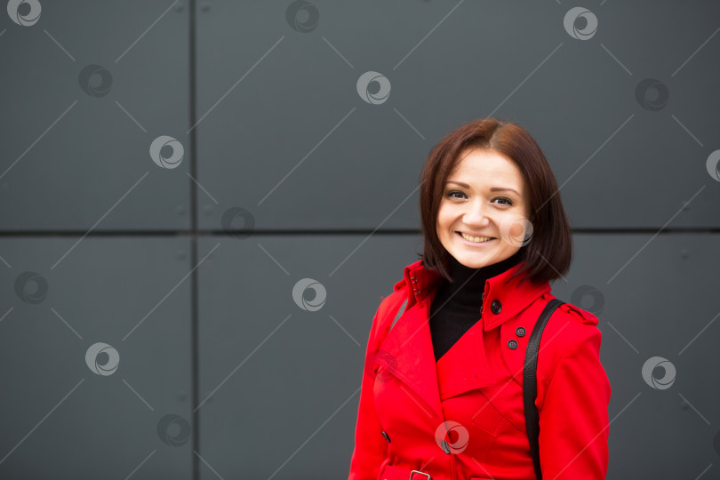 Скачать Молодая и счастливая кавказская женщина в красном плаще с черным кожаным рюкзаком на фоне серой стены. Образ жизни, эмоции, женственность, портрет фотосток Ozero
