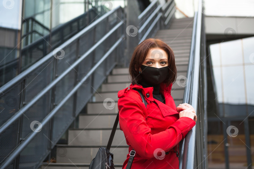 Скачать Молодая женщина в красном пальто в черной защитной медицинской маске возле стеклянного здания с лестницей. Эпидемия коронавируса, защитные меры, новая реальность фотосток Ozero
