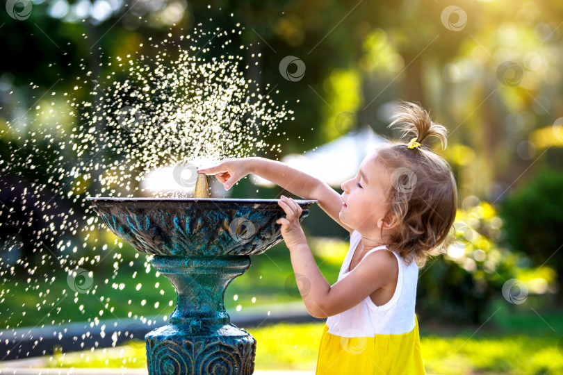 Скачать Забавная маленькая девочка играет с брызгами из фонтанчика с питьевой водой в парке. Детство, ребенок, летнее время, жара. Защита от солнечных лучей для детской кожи, фотосток Ozero