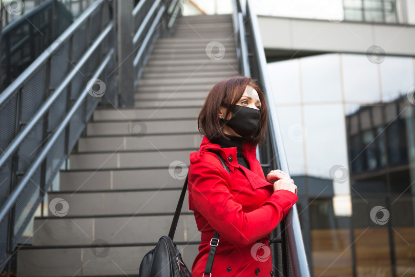 Скачать Молодая женщина в красном пальто в черной защитной медицинской маске возле стеклянного здания с лестницей. Эпидемия коронавируса, защитные меры, новая реальность фотосток Ozero