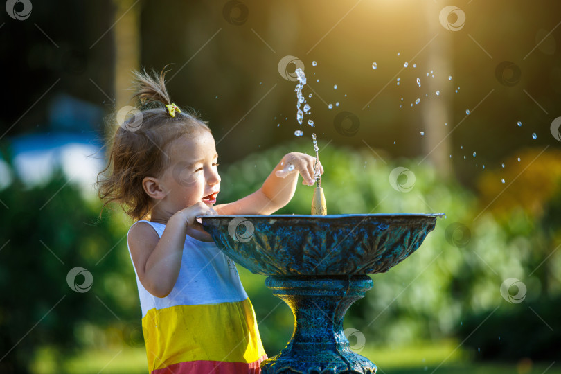 Скачать Забавная маленькая девочка играет с брызгами из фонтанчика с питьевой водой в парке. Детство, ребенок, летнее время, жара. Защита от солнечных лучей для детской кожи, фотосток Ozero