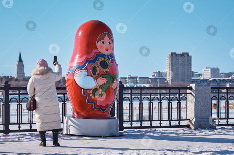 Скачать Большая матрешка на заснеженной зимней набережной реки Амур. Женщина в теплой одежде фотографирует матрешку на смартфон фотосток Ozero