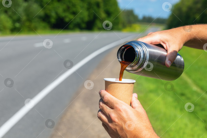 Скачать Путешественник наливает кофе из термоса в бумажный стаканчик на фоне дороги в солнечный день фотосток Ozero