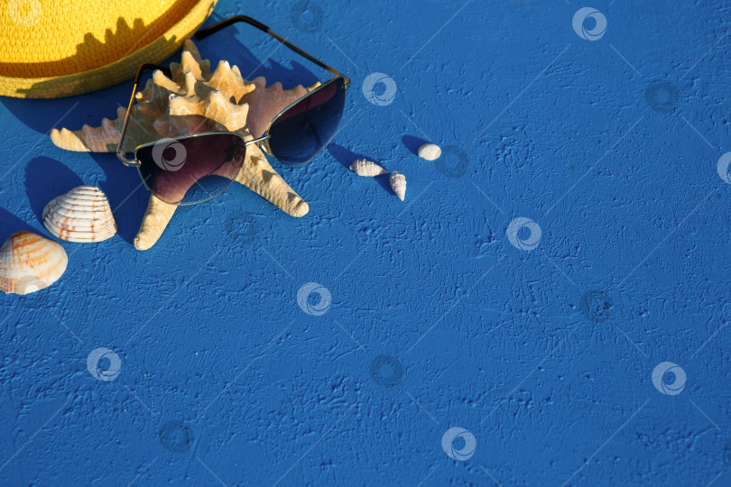 Скачать Рамка с пляжными аксессуарами на морскую тематику: желтая соломенная шляпа, солнцезащитные очки, морская звезда и ракушки на синем фоне. Концепция отпуска, морская прогулка, защита от ультрафиолета, плавание. Пространство для копирования. Плоский лист фотосток Ozero