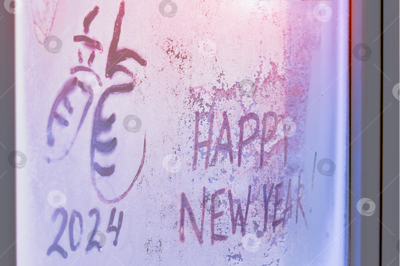Скачать Замерзшее окно с надписью на инее иероглифом дракон и поздравлением с Новым годом. Символ 2024 года. Солнечный свет проникает сквозь узор. Концепция Рождества и нового года. Избирательный фокус фотосток Ozero