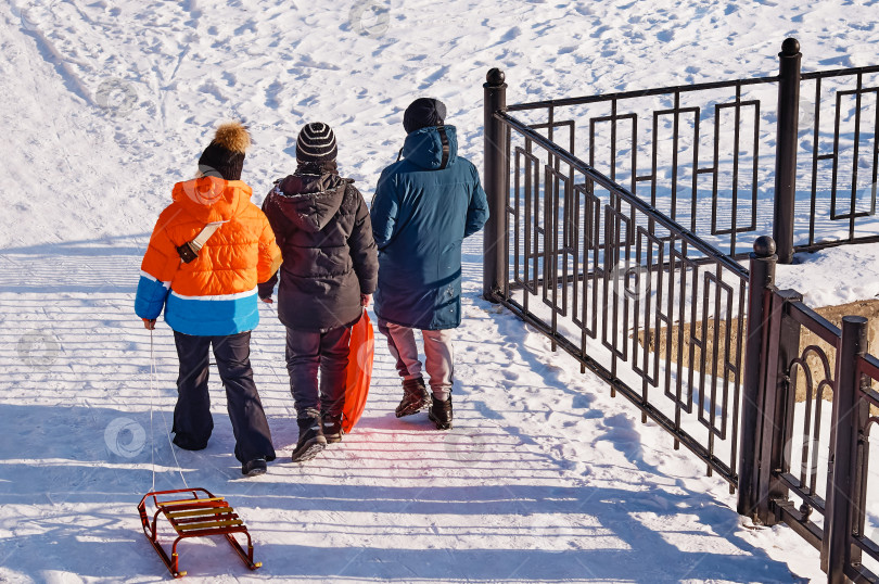 Скачать Группа подростков с санками идет по снегу, чтобы покататься на ледяной горке. Вид сзади на двух мальчиков и девочку в теплой одежде. Счастливые дети, играющие зимой в снегу на берегу реки. фотосток Ozero